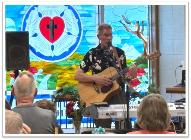 Musician playing acoustic guitar during a Lutheran worship service in front of a Luther Rose stained glass window